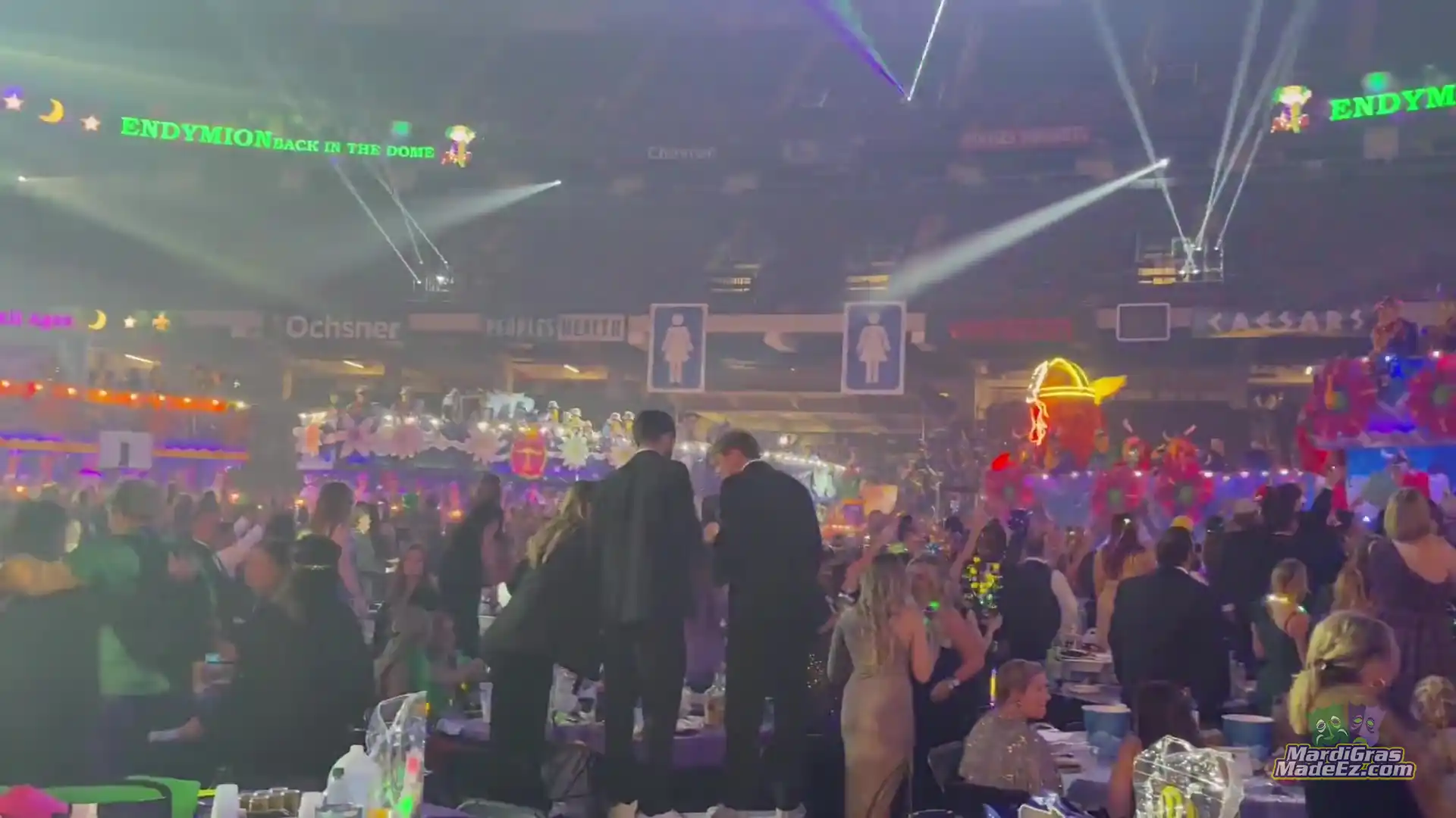 Guests in formal wear dancing between tables at a Mardi Gras ball under stadium lights